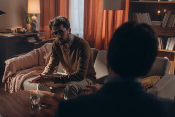 Young man in glasses sits on couch rubbing palms while sharing thoughts with psychologist