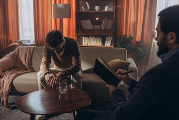 Psychologists writes in notebook while speaking with male patient during therapy session