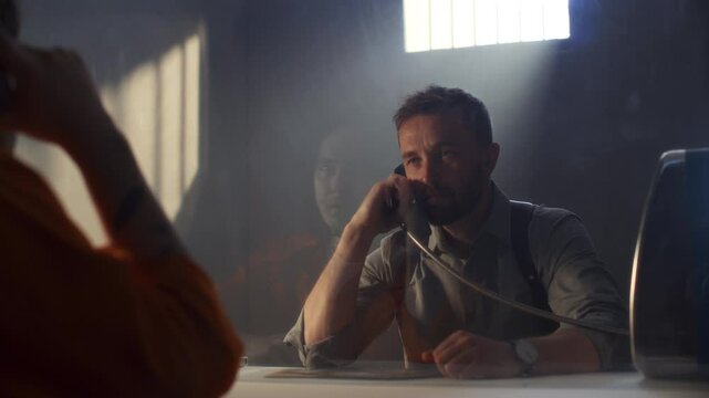 Investigator showing crime scene photo to prisoner and speaking on phone through glass barrier during interrogation in visitation room