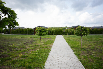 Brick pathway leading through an open field towards small trees and houses