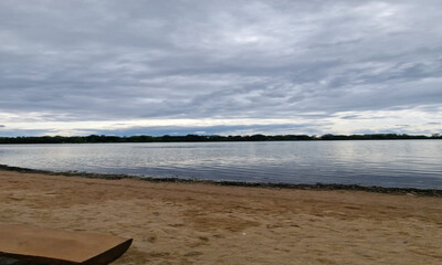 A tranquil lakeshore view with gentle ripples on the water, a sandy beach, and distant trees under...