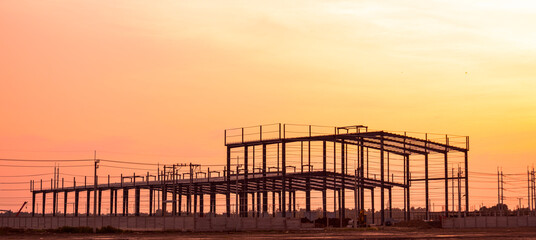 Fototapeta premium Silhouette metal structure of large warehouse factory building in construction site against orange sunset sky background, panoramic view