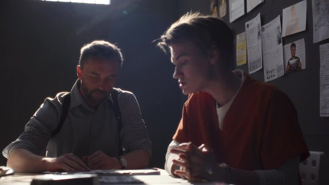 Young female inmate in orange uniform sitting across officer in prison room, denying involvement in crime as reviewing photos of criminal evidence during tense interrogation