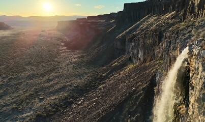 Aerial view of a waterfall cascading down a rocky cliffside with a vast desert landscape at sunset