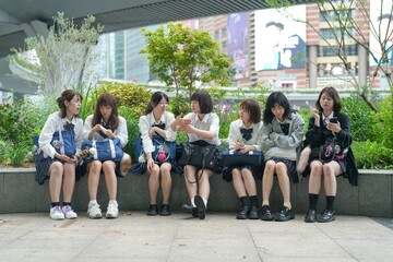 In early summer in Shanghai, near The Bund, Japanese high school girls in uniforms sit at a street corner, chatting and laughing, their smiles adding a touch of youth to the vibrant city atmosphere.