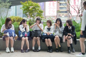 Fototapeta premium In early summer in Shanghai, near The Bund, Japanese high school girls in uniforms sit at a street corner, chatting and laughing, their smiles adding a touch of youth to the vibrant city atmosphere.