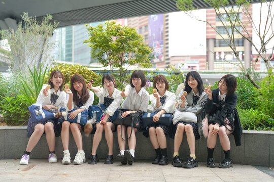 In early summer in Shanghai, near The Bund, Japanese high school girls in uniforms sit at a street corner, holding Chinese snacks and posing for a commemorative photo with cheerful expressions.
