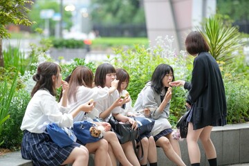In early summer in Shanghai, near The Bund, Japanese high school girls in uniforms sit at a street corner, enjoying Chinese sweets, their cheerful expressions adding charm to the vibrant city scene.
