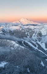 Aerial view of a snow covered mountain range with evergreen trees and a pink and orange sky at sunset