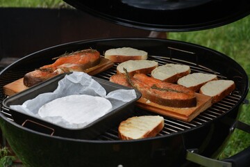 Full view of black charcoal grill standing on green grass, loaded with salmon, cheese, and bread