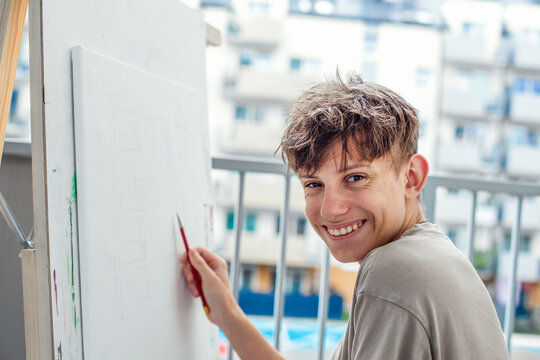 Cheerful, cute teenager at easel learning to draw with pencil on easel, smiling sincerely, looking at camera