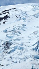 A snowy mountain slope with visible crevasses and rocky outcrops under a cloudy sky in winter