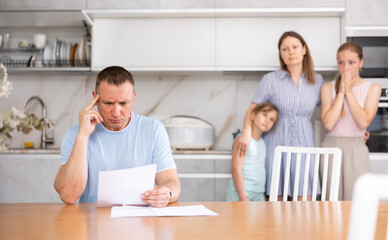 Fototapeta premium Saddened head of family is sitting in kitchen and reading final documents of family business. Wife and children are standing behind man and waiting for news from Dad