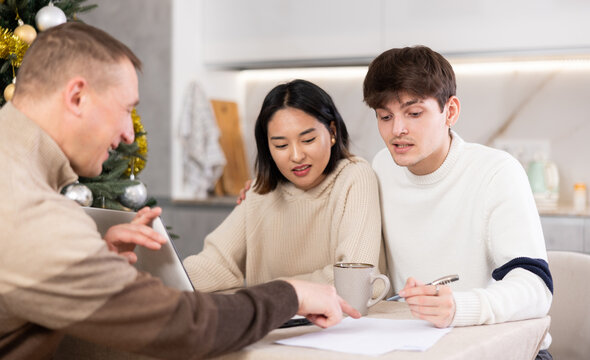 Male realtor discusses the terms of contract with couple in office of real estate agency, offering various accommodation options against backdrop of a Christmas tree