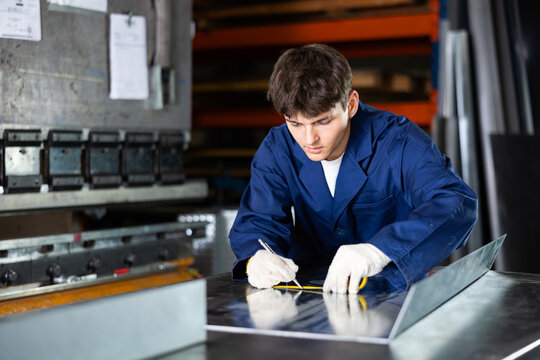 Young guy, skilled worker in metal workshop, focusing on accurately marking steel sheet with tape measure, preparing workpiece for further cutting, drilling, or assembling components..