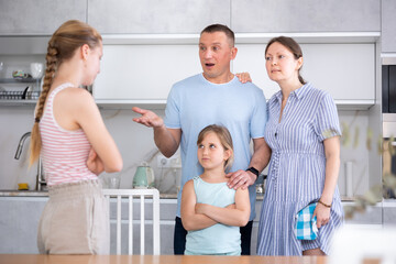 Teenage girl with long pigtail standing in home kitchen, listening to disapproving words of father and mother. Concept of tension and disagreement arising between generations..