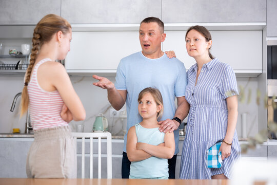 Teenage girl with long pigtail standing in home kitchen, listening to disapproving words of father and mother. Concept of tension and disagreement arising between generations..
