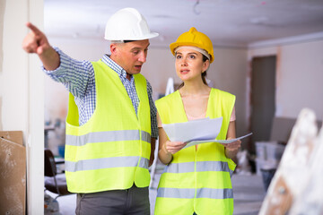 Portrait of woman architect and foreman holding blueprints discussing working process and gesturing in house being renovated