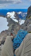 A person sitting on a rocky mountain ledge with a blue climbing rope and snowy mountain views behind
