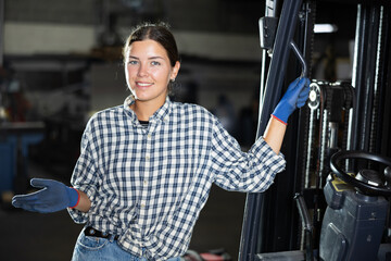Young female worker in uniform posing by forklift in metallurgical workshop