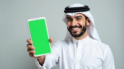 young arabian muslim bearded man wearing national clothes, smiling looking at camera and showing empty green screen mobile phone on white light grey background. mockup for advertising