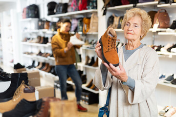 Fototapeta premium Old lady standing in salesroom of shoe store and choosing new boots. Younger woman shopping in background.
