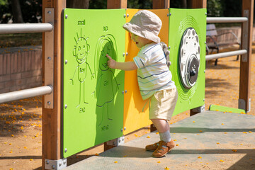 Happy emotional baby boy wearing summer clothes explores a playground. He is interacting with a green activity panel featuring simple drawings of a boy and girl designed for sensory play. 