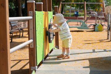 A baby boy wearing a sun hat, striped shirt, and shorts explores a playground. He is interacting with a green activity panel designed for sensory play.  Sunny summer weather