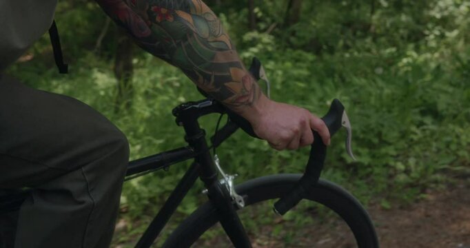 A close-up of tattooed forearms tightly gripping a black bicycle handlebars. The natural forest light and green surroundings highlight the raw, grounded energy of an offline journey. High quality 4k