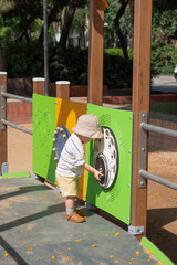Baby playing outdoors on little kids playground. The child using both hands to explore the panel witch features musical symbols and a circular, interactive component resembling a control dial. 