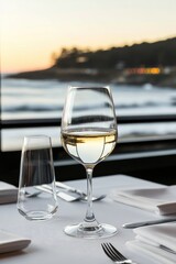 Obraz premium A glass of white wine sits on a table set for two, overlooking a sun-drenched ocean vista at sunset
