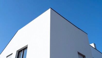 青空を背景に、低い視点から捉えたモダンでミニマリストな白い壁と鋭いエッジが特徴の住宅が、コントラストの強い光により洗練された姿を見せる

