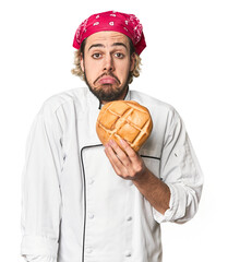 Young chef with artisan bread in studio shrugs shoulders and open eyes confused.