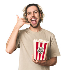 Young man holding popcorn for a movie showing a mobile phone call gesture with fingers.