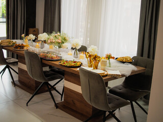 Elegant table setup for a celebration or private event, featuring a variety of delicious foods, white floral arrangements, golden utensils, and modern interior design. Natural daylight highlights the 