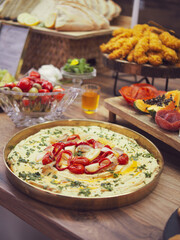 A vibrant and inviting food spread arranged on a rustic wooden table. The centerpiece is a generous bowl of creamy hummus, beautifully garnished with herbs, red peppers, and olive oil. Surrounding it 