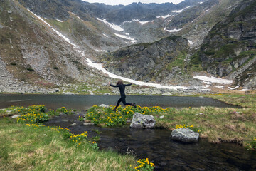 Hiker celebrating freedom by jumping across alpine brook, golden flowers framing scenic glacial valley with steep rocky slopes and snowfields
