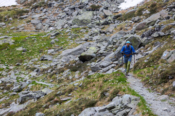 Mountain hiker with backpack and trekking poles navigating rocky alpine trail through rugged terrain filled with boulders and scattered stones