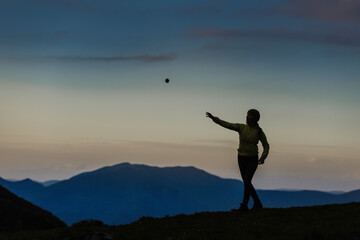 Adventure enthusiast enjoying playful moment at sunset, throwing and catching ball while standing on scenic mountain overlook at dusk.
