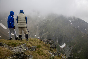 Two hikers with trekking poles standing on misty mountain ridge, contemplating dramatic alpine valley with snow patches and foggy weather.