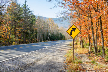Yellow traffic sign warning drivers agaisnt a dangerous curve along a mountain road on a sunny autumn day
