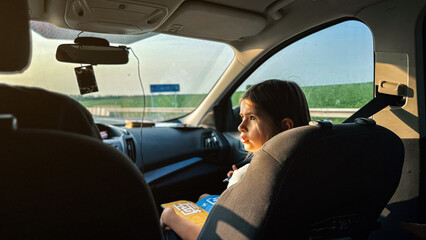 Girl child 8 -9 years sitting in the seat of a car during road trip, holding an activity book and looking out into the sunset light with curiosity, symbolizing childhood and travel, childhood concept