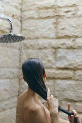 Woman washing long dark hair under outdoor shower with stone wall background, natural skin tones, relaxed posture, and fresh water droplets.