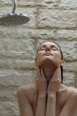 woman showering under rain showerhead with relaxing expression, stone wall background, wet skin, wellness ritual, natural light