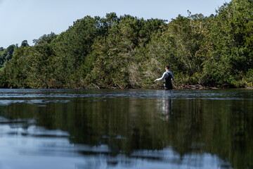 A young man immersed waist-deep in a tranquil river focuses on fly fishing, surrounded by nature and soft light.