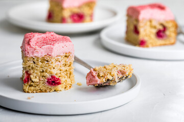 A piece of cake with pink frosting and raspberries on a white plate, with a fork holding a bite-sized piece.