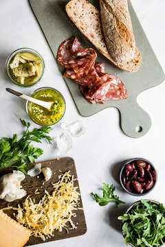 An assortment of ingredients including a baguette, sliced salami, artichokes, grated cheese, olives, and arugula are arranged on a cutting board and table.