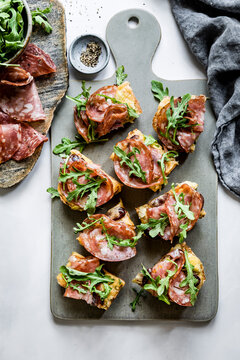An assortment of open-faced sandwiches with salami and arugula on a serving board, accompanied by a bowl of arugula and a dish of pepper.