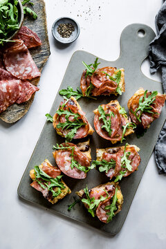 An assortment of open-faced sandwiches with salami and arugula on a serving board, accompanied by a bowl of arugula and a dish of black pepper on a white background.