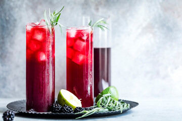 Two tall glasses of red berry cocktail with ice, garnished with herbs, served on a tray with lime and blackberries.